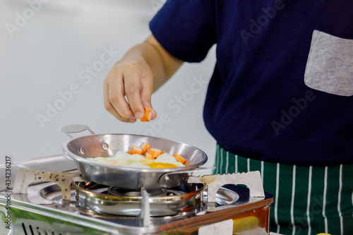 Smart kid on black shirt of modern family carefully pouring ingredients into flying pan for cooking easy breakfast as learning from mom in home kitchen