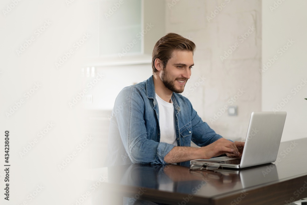 Young casual man working on laptop online, sitting at table in kitchen, looking at computer screen Young casual man working on laptop online, sitting at table in kitchen, looking at computer screen