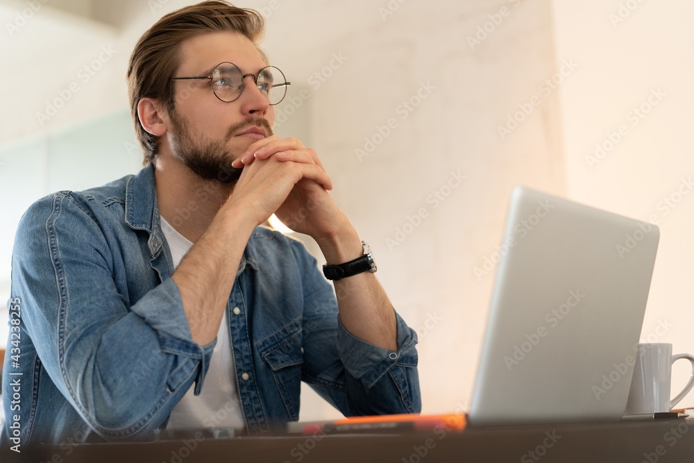 Young casual man working on laptop online, sitting at table in kitchen, looking at computer screen Young casual man working on laptop online, sitting at table in kitchen, looking at computer screen