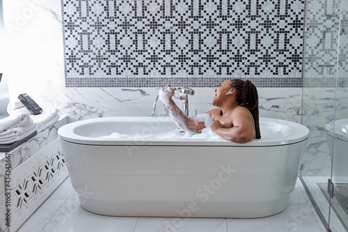Cute african woman taking bath with bubbles in bathroom interior, singing in shower handle, have fun. Lifestyle wellness concept. Treating yourself and wellbeing.