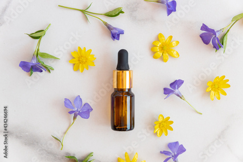 Blank amber glass essential oil bottle with pipette on marble background decorated blooming wild flowers. Skin care concept with natural cosmetics.