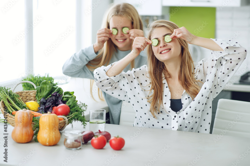 Young and happy woman with her daughter eating a healthy salad sits on a table with green fresh ingredients indoors. High quality photo. Young and happy woman with her daughter eating a healthy salad sits on a table with green fresh ingredients indoors. High quality photo.