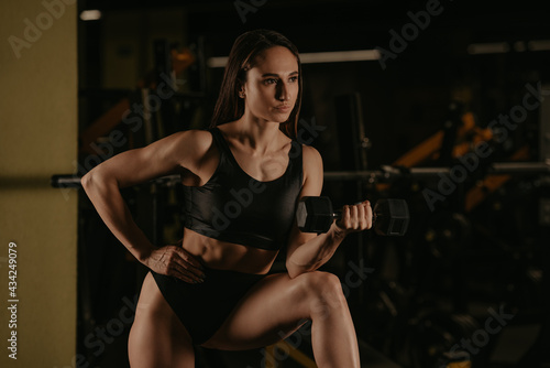 A sporty woman is doing dumbbell curls with her elbow on a knee in a gym. A muscular brunette girl wears a black top and high waist short shorts.