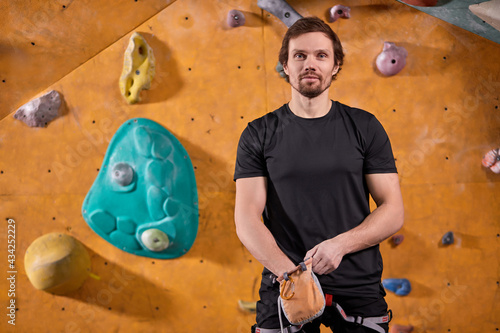 Portrait Of Handsome Caucasian Guy Sportsman Climber Is Going To Climb Artificial Rock Wall, Look At Camera Smiling, Extreme Sports And Bouldering Concept. Copy Space