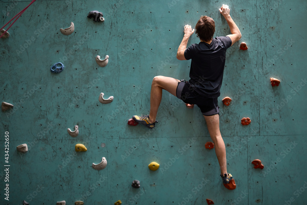 active young caucasian man on rock wall in sport center, rock climbing. Sportive male guy in black sportswear engaged in sport, fitness, lead healthy lifestyle. Rear view - obrazy, fototapety, plakaty 