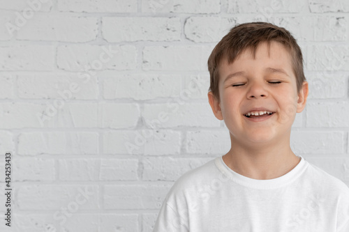 A little boy in a white T-shirt on a light background laughs, closing his eyes. Free space for text placement