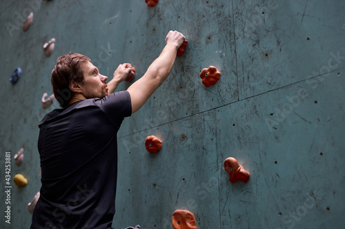 Caucasian strong sportsman climber moving up on steep rock, climbing on artificial wall indoors, side view, copy space. Extreme sports and bouldering concept