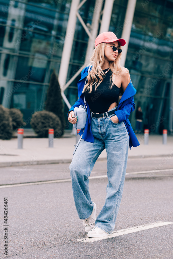 Young sexy blonde hipster woman posing on the street. Wearing blue stylish jacket, jeans and baseball hat and sunglasses.