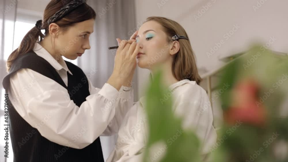 Female visagiste doing makeup for model. Low angle of professional makeup artist applying blue eyeshadows on eyelids of young client with charming appearance in beauty studio Female visagiste doing makeup for model. Low angle of professional makeup artist applying blue eyeshadows on eyelids of young client with charming appearance in beauty studio