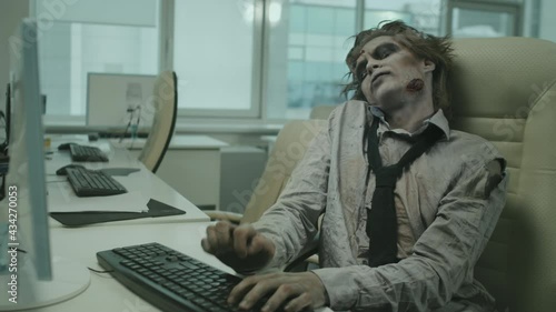Medium shot of zombie office worker sitting at desk and smashing keyboard with his hands while trying to type