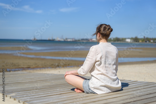 Woman in linen clothes doing yoga lotus pose on beach at sunny day. Wellbeing, physical and mental health. Looking to water