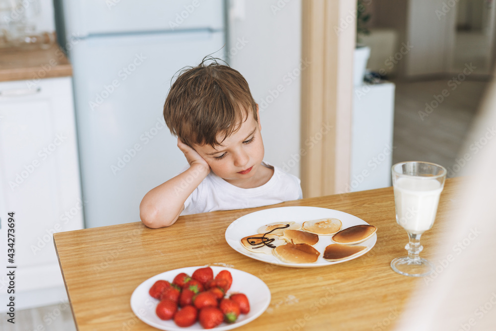 Cute toddler boy sitting with puncakes with berries in kitchen at the home. Child doesn't want to eat Cute toddler boy sitting with puncakes with berries in kitchen at the home. Child doesn't want to eat
