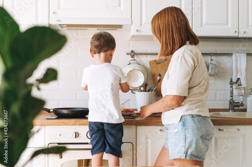 Cute toddler boy with his mother cooking breakfast with puncakes together in bright kitchen at home photo