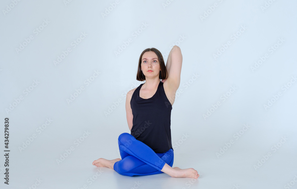 Fototapeta premium Cute female doing yoga exercise cow face pose in blue sports leggings and black top, on white background isolated on cyclorama. Copy space text. Gomukhasana.