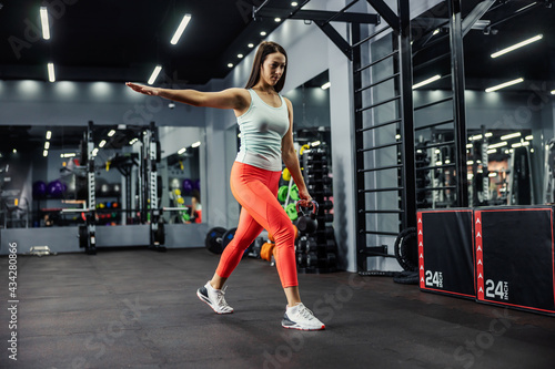 A dynamic young girl in red leggings and a mint T-shirt is doing a step forward in the gym on a black background. She raised up one hand horizontally and she holds a kettle-bell in her other hand