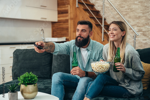 Fototapeta Naklejka Na Ścianę i Meble -  Young beautiful couple drinking beer, eating popcorn and watching sport game on a television at home