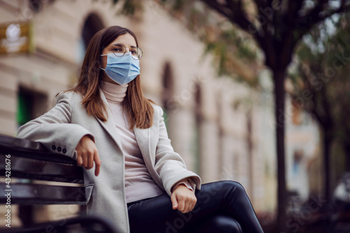 Female influencer with face mask sitting on the bench outside during corona virus or air pollution. © dusanpetkovic1