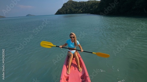 Pretty sportive blonde lady in stylish swimsuit sails on pink plastic canoe along azure ocean bay at exotic resort.