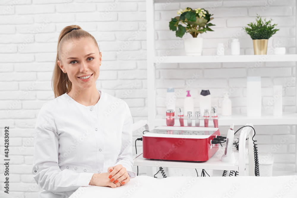 Front view portrait of smiling young beautiful woman sitting in clinic near special beauty equipments and waiting for clients. Concept of preparing for beauty procedures with modern cosmetics. Front view portrait of smiling young beautiful woman sitting in clinic near special beauty equipments and waiting for clients. Concept of preparing for beauty procedures with modern cosmetics.