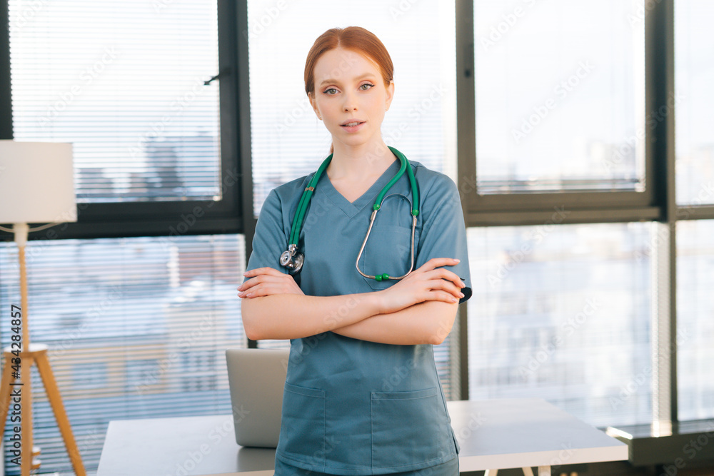 Portrait of happy smiling young female doctor wearing blue green uniform and stethoscope looking at camera. cheerful redhead woman physician posing in hospital office near window. Portrait of happy smiling young female doctor wearing blue green uniform and stethoscope looking at camera. cheerful redhead woman physician posing in hospital office near window.