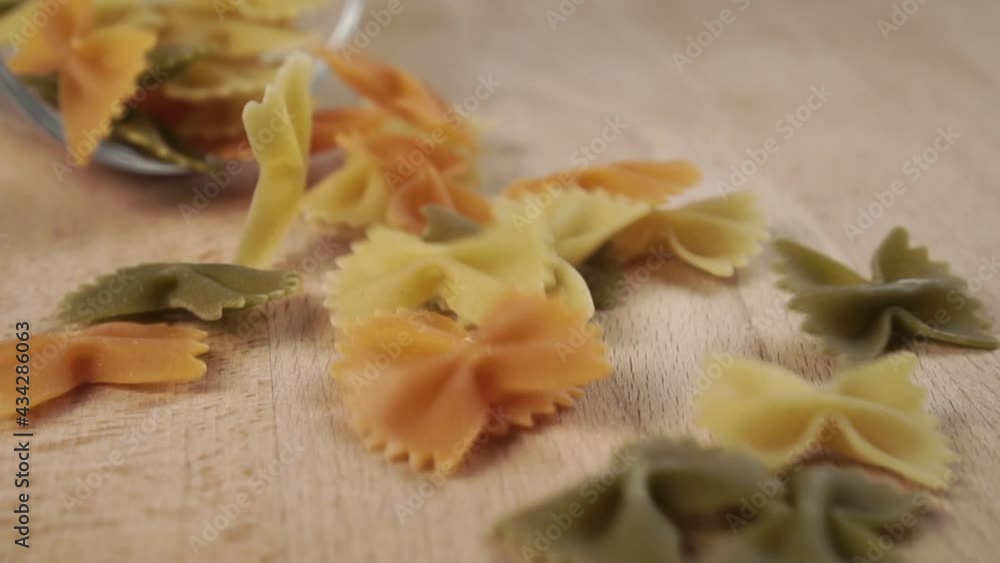 Colored uncooked butterfly pasta falling in slow motion from a transparent glass cup. On a wooden cutting board. Raw farfalle. Close-up Colored uncooked butterfly pasta falling in slow motion from a transparent glass cup. On a wooden cutting board. Raw farfalle. Close-up