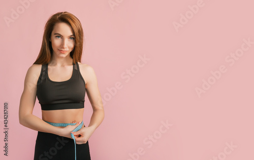 Fitness girl smiles and measures the waist in black sportswear on a pink background. Slim woman with a beautiful athletic body and tanned skin © Daria Lukoiko