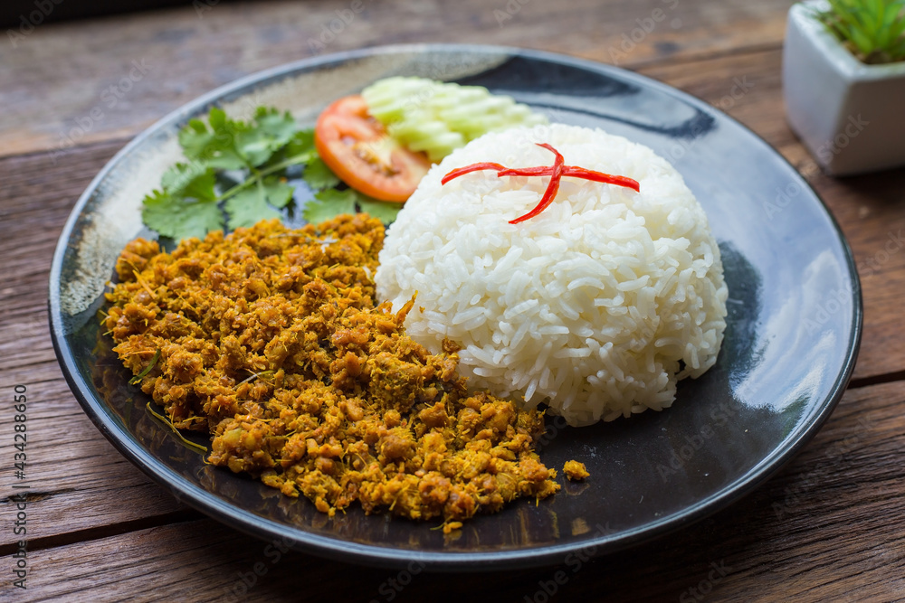 Stir fried pork with yellow curry paste over rice is on a black plate and a brown wood as background. It is a southern local food of Thailand. Stir fried pork with yellow curry paste over rice is on a black plate and a brown wood as background. It is a southern local food of Thailand.
