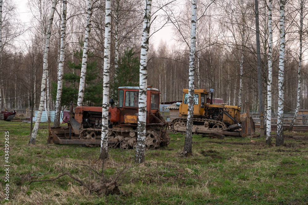 Old red and yellow tractors stand among the trees in the open air on a driverless farm. Agricultural machinery Old red and yellow tractors stand among the trees in the open air on a driverless farm. Agricultural machinery