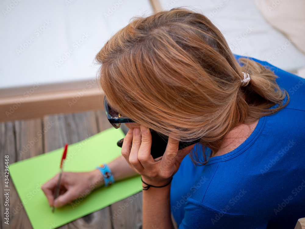Fototapeta premium Close-up of happy blonde woman in sunglasses talking on the phone and writing on a paper