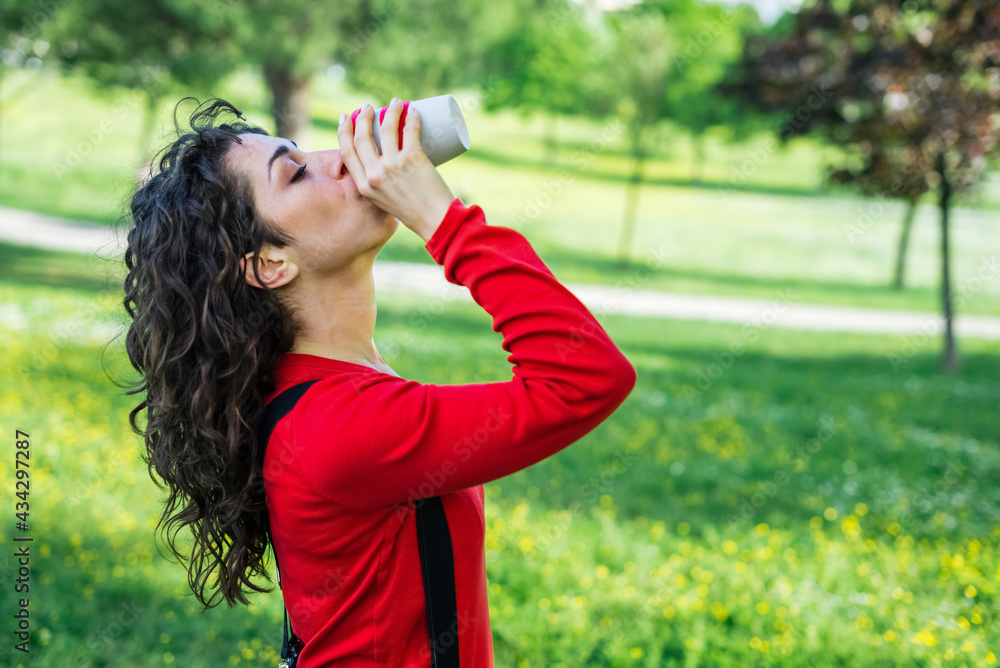 Beautiful woman drinking coffee to go in a park outdoor in summer. Portrait of young adult girl outside sipping hot cappuccino and looking away. Pretty female with reusable modern cup of drink. Beautiful woman drinking coffee to go in a park outdoor in summer. Portrait of young adult girl outside sipping hot cappuccino and looking away. Pretty female with reusable modern cup of drink.