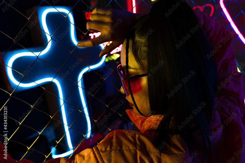 stylish young asian woman in sunglasses posing near neon sign and metallic fence