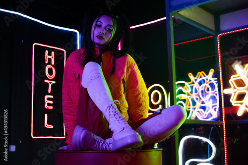 young asian woman sitting near neon sign with hotel lettering outside