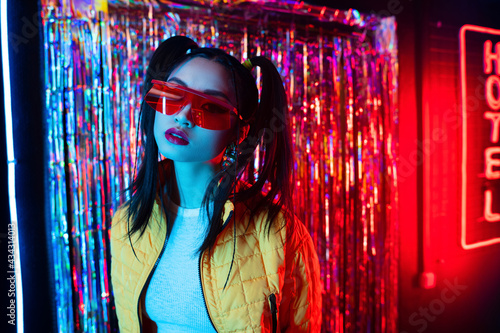 young asian woman in sunglasses looking at camera near red neon hotel sign photo