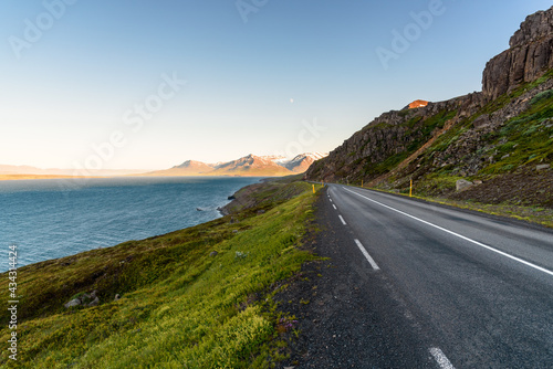 Deserted road along a rugged coast of a fjord under clear sky in summer. Some snow capped coastal mountains warmly lit by a setting sun are visible on horizon. Northern Iceland.