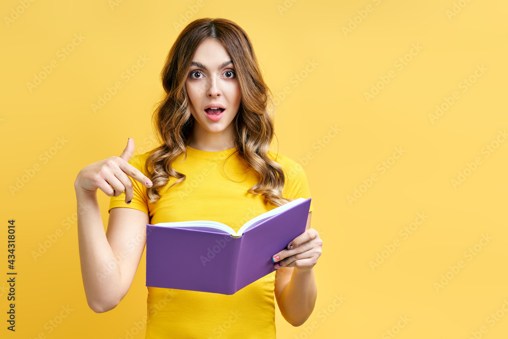 Portrait of amazed surprised woman pointing on book with copy space on yellow background Portrait of amazed surprised woman pointing on book with copy space on yellow background