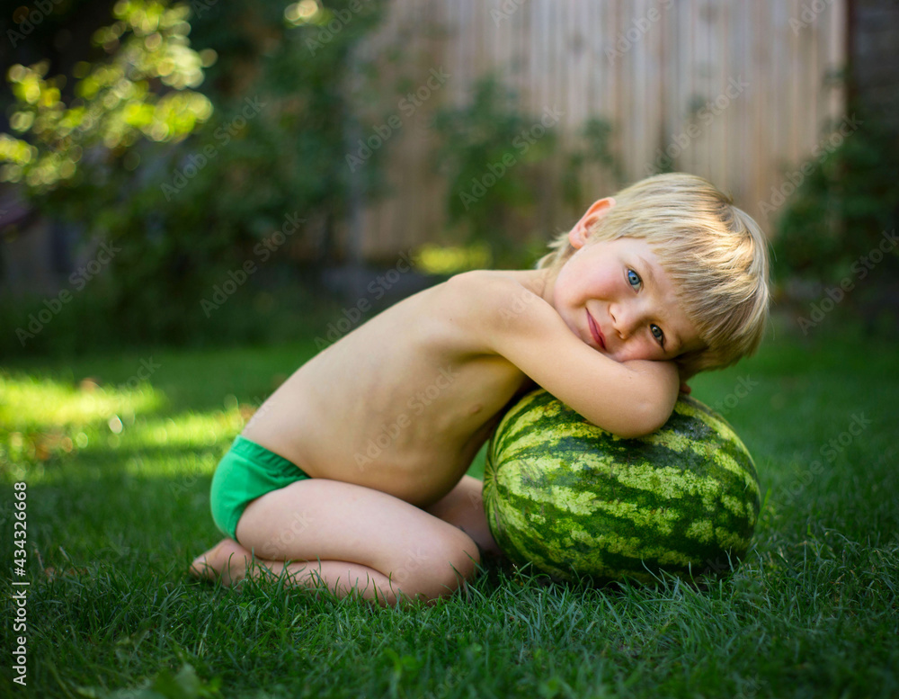 On a hot summer day, a cute barefoot toddler boy without clothes sits on the grass next to a large striped watermelon. Waiting for an appetizing delicious dessert. Cheerful carefree childhood On a hot summer day, a cute barefoot toddler boy without clothes sits on the grass next to a large striped watermelon. Waiting for an appetizing delicious dessert. Cheerful carefree childhood