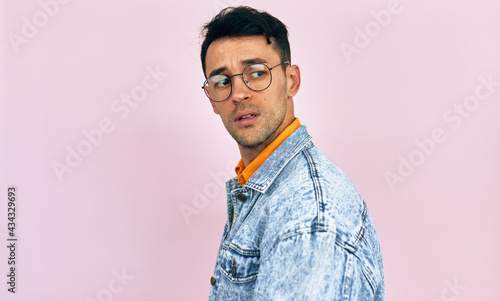 Handsome young man has uncertain exression on his face, isolated on pink studio background. Male wearing stylish casual clothes, transparent eyeglasses has doubtful expression. photo