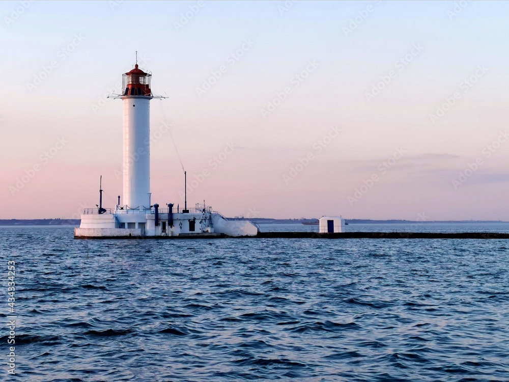  White lighthouse on the background of the beach and the water of the Black Sea near the coast of Odessa. The color of the water is blue, gentle sunset