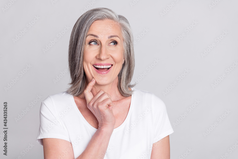 Photo of confident impressed mature lady wear white clothes finger chin looking empty space smiling isolated grey color background Photo of confident impressed mature lady wear white clothes finger chin looking empty space smiling isolated grey color background
