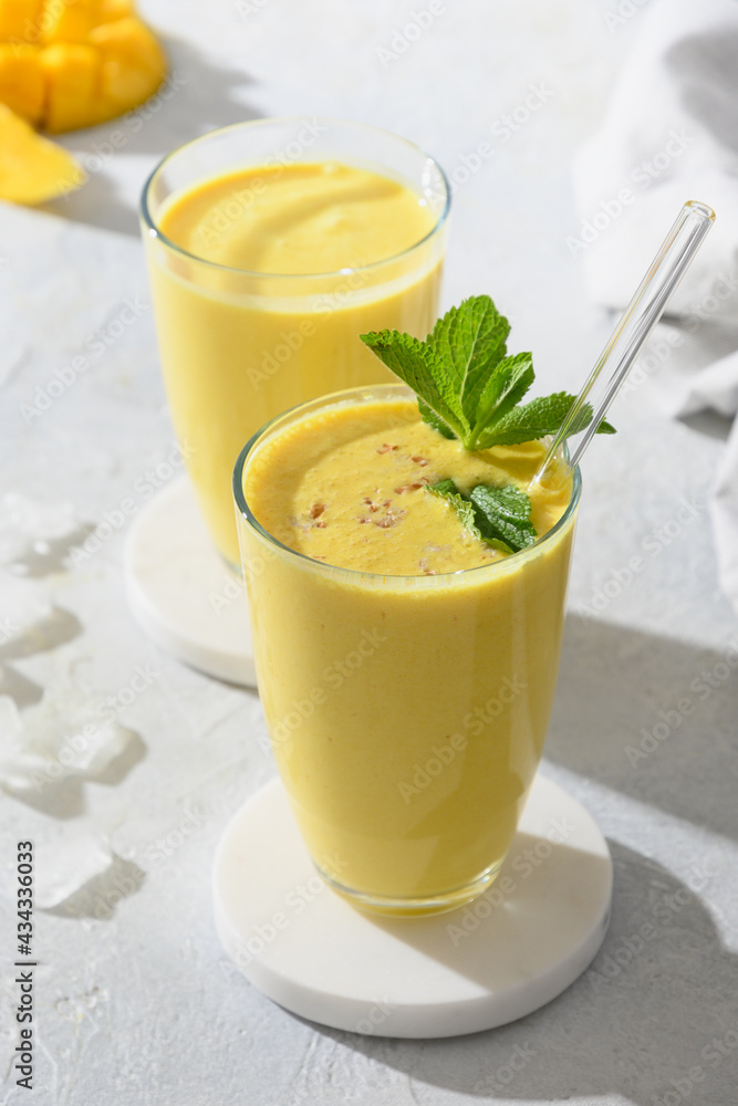 Two mango lassi or smoothies on a gray background with sunny shadow. Indian healthy ayurvedic cold drink with mango, yogurt, water, spices, fruits and ice. Close up.