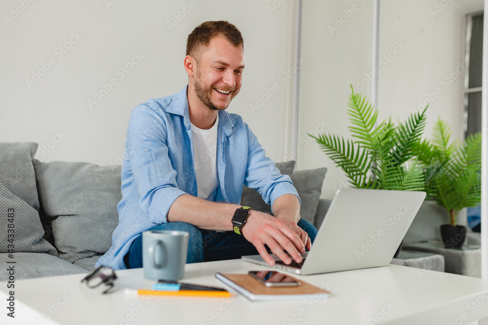 handsome smiling man sitting on sofa drinking tea at home at table working online on laptop handsome smiling man sitting on sofa drinking tea at home at table working online on laptop