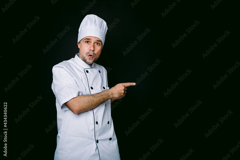 Chef cook wearing jacket and kitchen hat pointing right on black background Chef cook wearing jacket and kitchen hat pointing right on black background