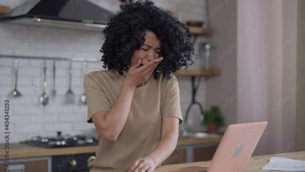 Tired young African American woman yawning looking at laptop screen in kitchen. Portrait of exhausted overworked slim beautiful manager in home office indoors. Tiredness and exhaustion Tired young African American woman yawning looking at laptop screen in kitchen. Portrait of exhausted overworked slim beautiful manager in home office indoors. Tiredness and exhaustion