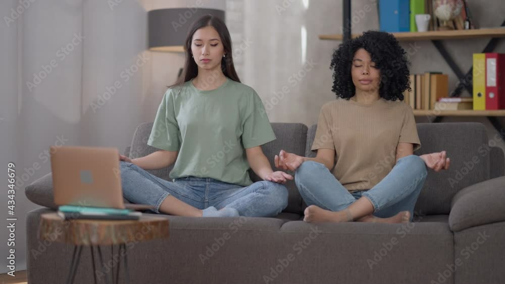 Wide shot of two calm confident slim young women sitting on couch in lotus position with eyes closed. Beautiful Asian and African American friends meditating in living room indoors. Meditation yoga Wide shot of two calm confident slim young women sitting on couch in lotus position with eyes closed. Beautiful Asian and African American friends meditating in living room indoors. Meditation yoga