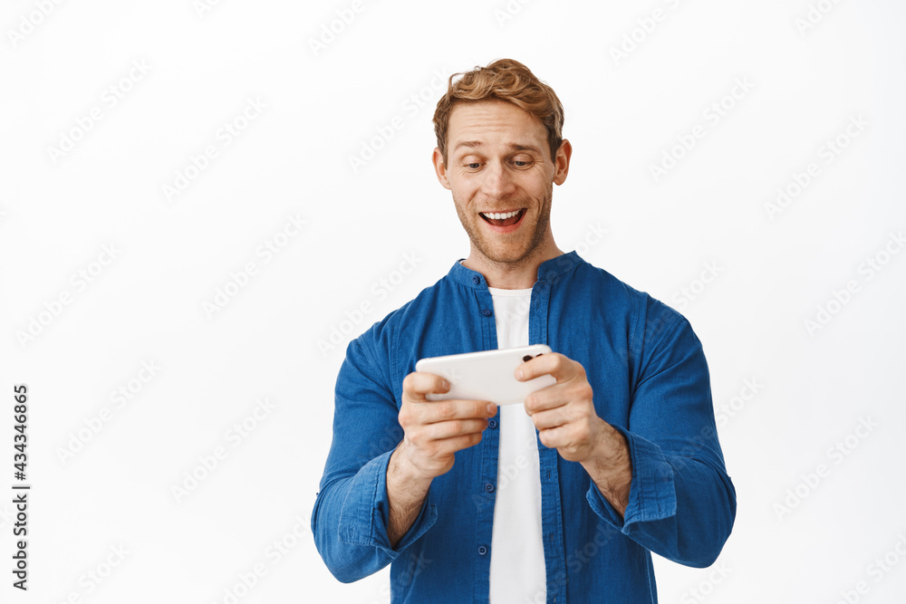 Happy redhead guy watching videos on smartphone, looking at mobile phone screen while playing video game app, smiling amused, standing over white background Happy redhead guy watching videos on smartphone, looking at mobile phone screen while playing video game app, smiling amused, standing over white background