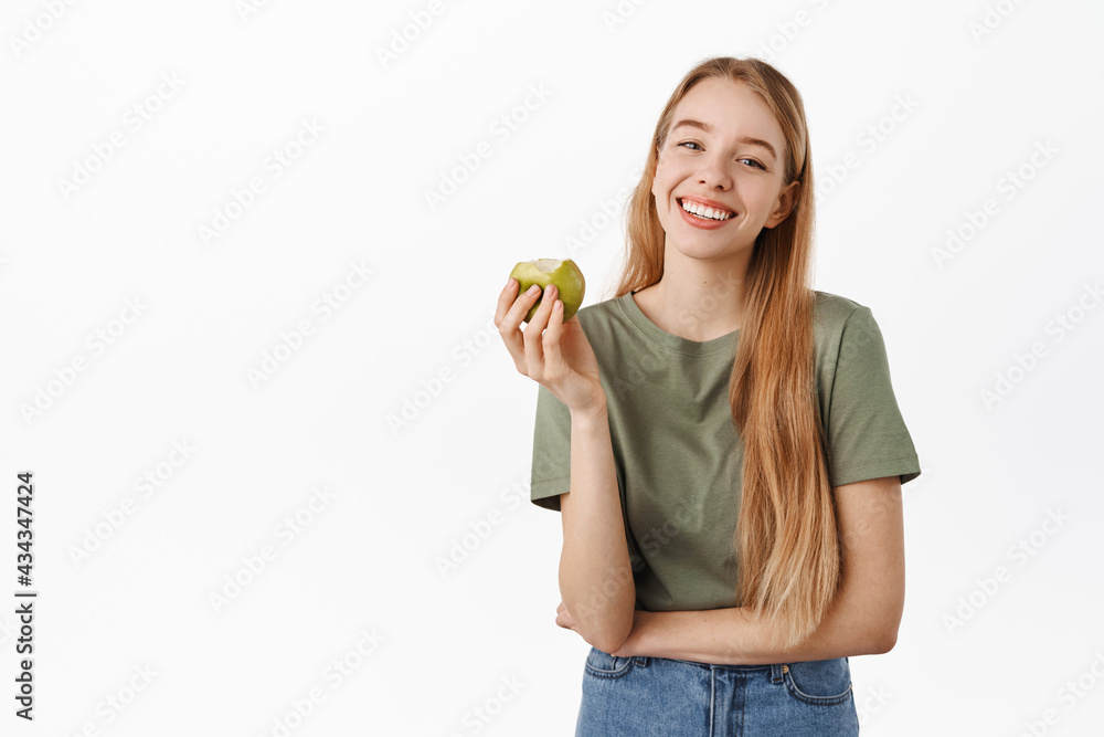 Dentist, stomatology concept. Smiling young woman holding green apply and showing perfect white smile teeth, standing happy and looking at camera over white background