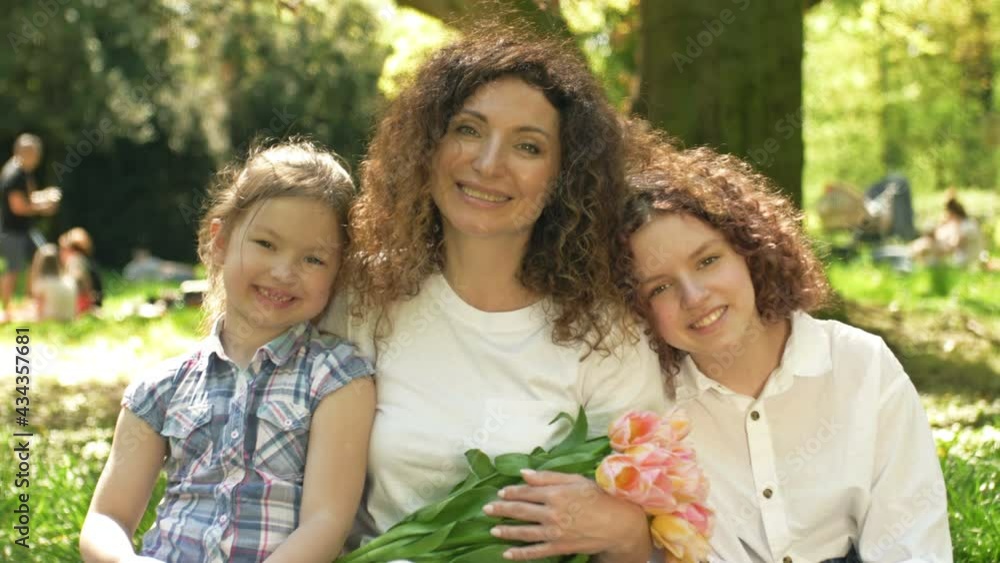 Two girls of different ages hug their mother. The woman is holding a beautiful bouquet of tulips. Against the background of a spring park. Mothers Day. Two girls of different ages hug their mother. The woman is holding a beautiful bouquet of tulips. Against the background of a spring park. Mothers Day.