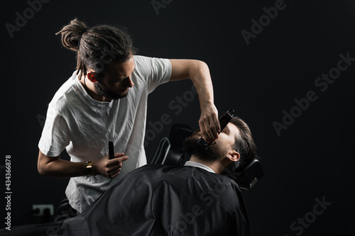 Handsome bearded man has hair and beard cut in barbershop. Dreadlock barber cuts client's hair