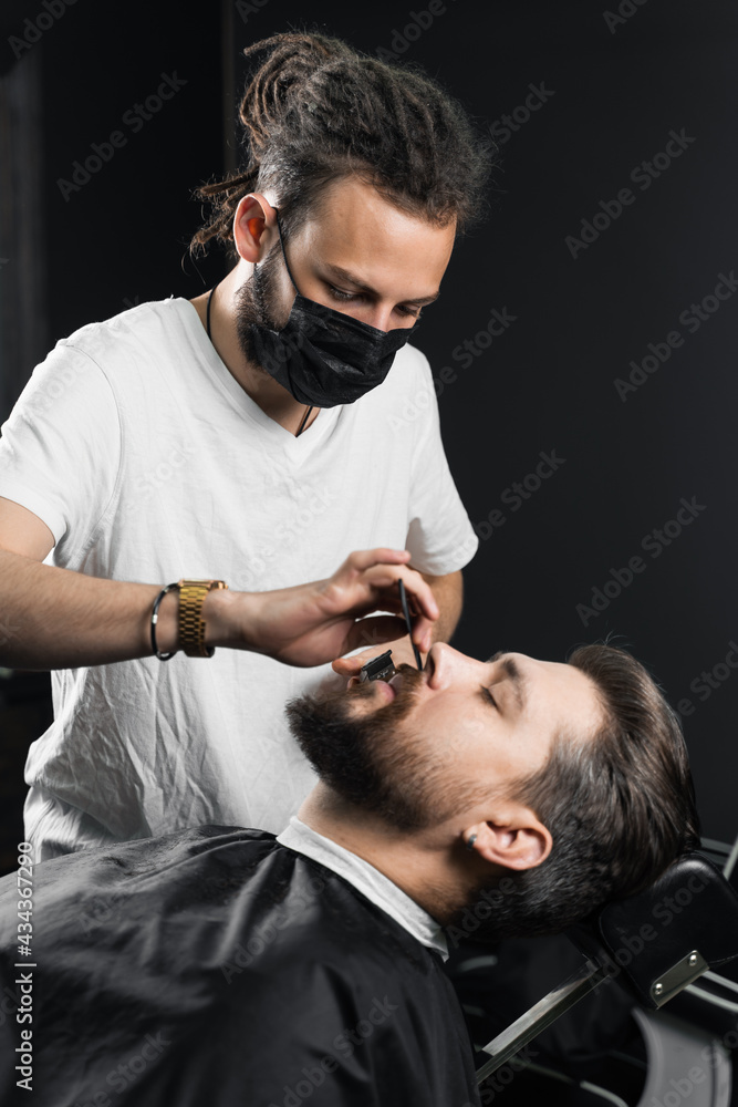 Fototapeta premium Mustache shaving in barbershop. Barber with dreadlocks in black medical mask trim beard of handsome man at quarantine coronavirus covid-19.