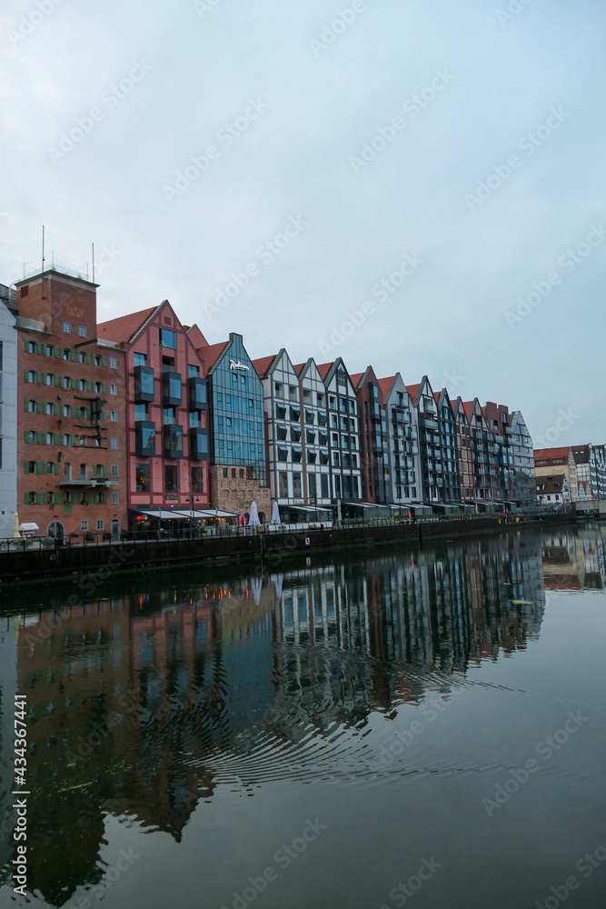 Fototapeta premium A panoramic view on the shores of Martwa Wisla flowing through Gdansk in Poland. The buildings reflect in the calm surface of the river. New architecture meeting with medieval constructions. Overcast
